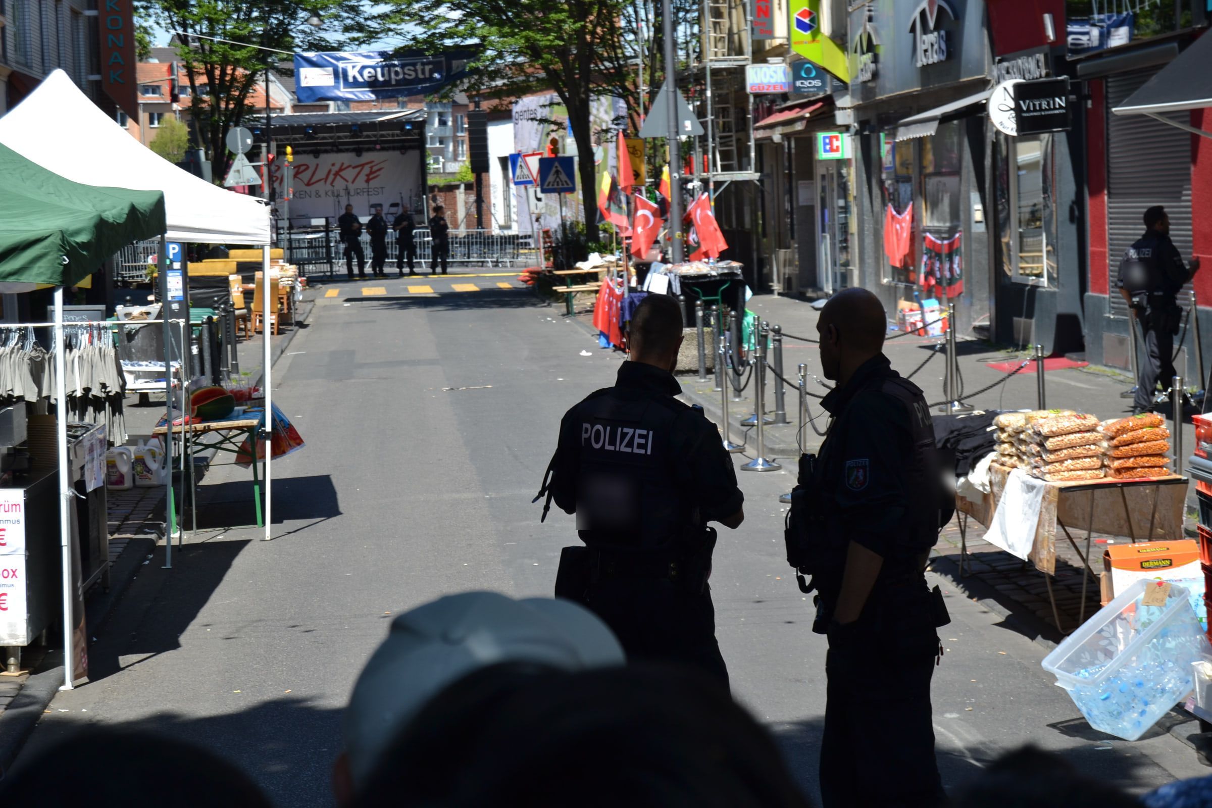 Blick die Keupstraße hinunter während des Birlikte-Fests: Marktstände, türkische Fahnen, ein Banner „Keupstr.“ über der Straße, dahinter die Bühne mit dem Schriftzug Birlikte; im Vordergrund zwei Polizisten der NRW-Polizei mit dem Rücken zur Kamera.