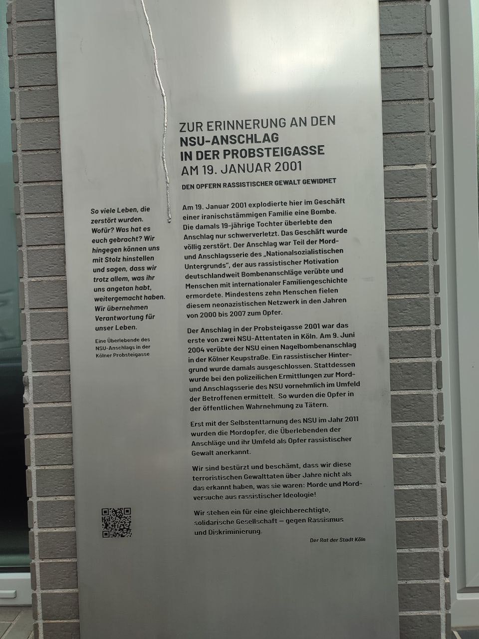 Eine Stele aus gebürstetem Stahl zwischen grauen Ziegelpfeilern mit eingraviertem Text: „Zur Erinnerung an den NSU-Anschlag in der Probsteigasse am 19. Januar 2001. Den Opfern rassistischer Gewalt gewidmet“, gezeichnet vom Rat der Stadt Köln; links das Zitat einer Überlebenden.